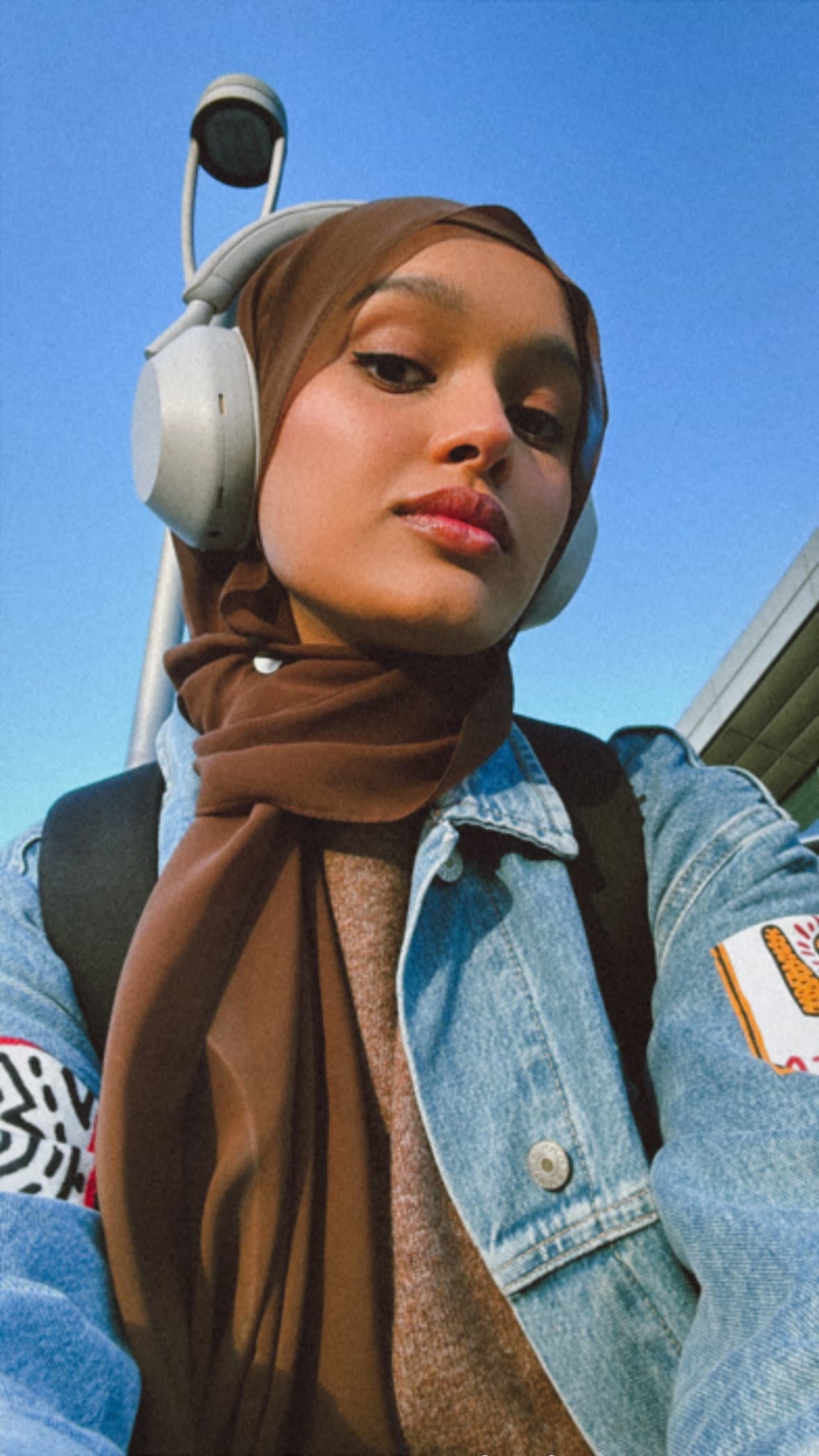 Woman wearing a brown hijab and denim jacket with white headphones against blue sky.