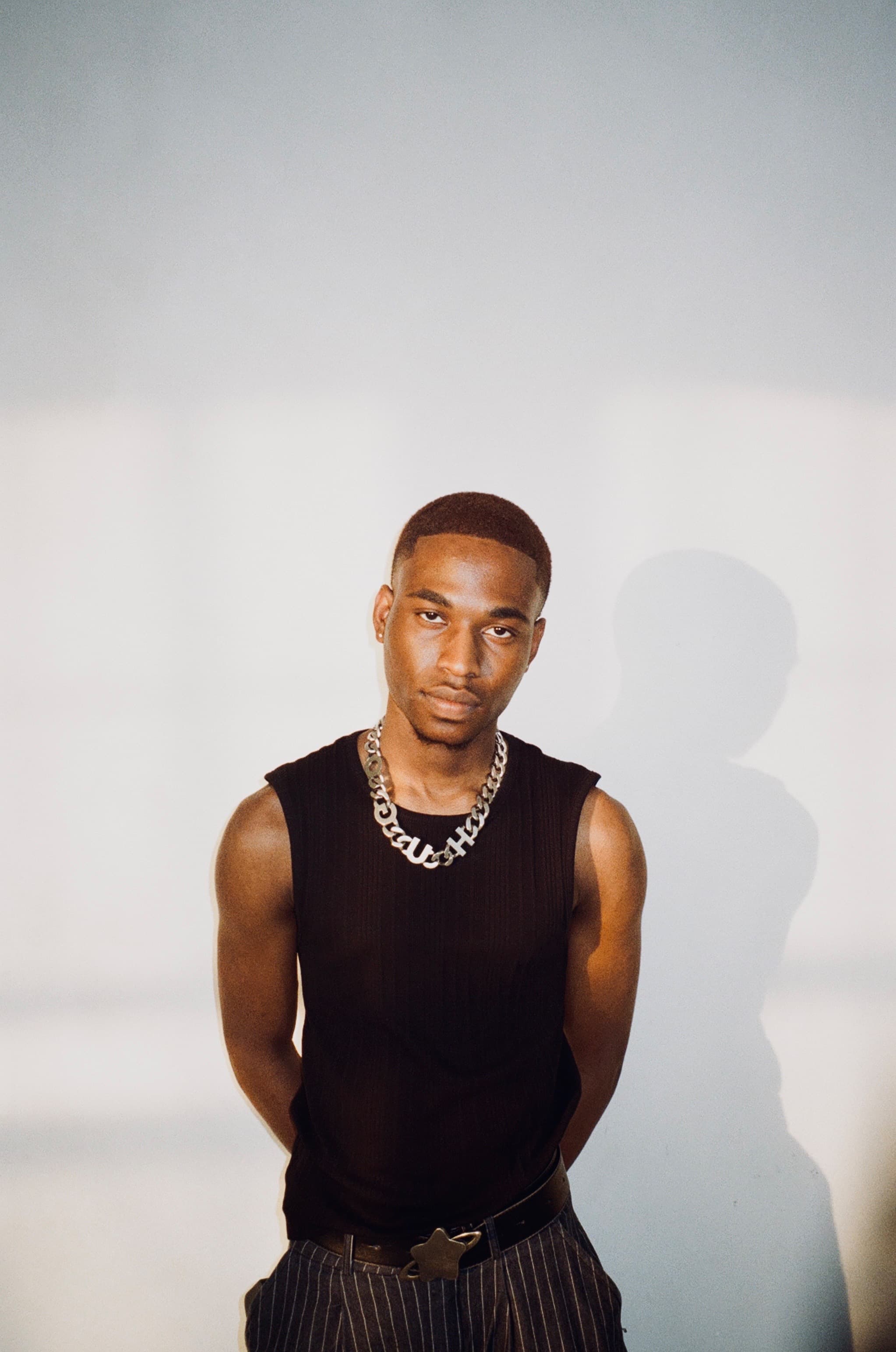 Young Black man in black sleeveless top and chunky silver chain against a white wall.